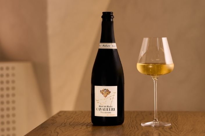 Bottiglia di Cavalleri Franciacorta DOCG Blanc de Blancs Nature accanto a un calice di spumante dorato su un tavolo di legno.