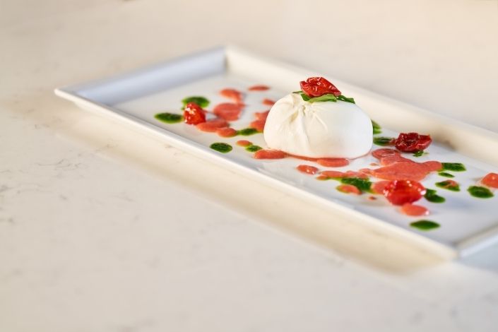 Piatto gourmet con burrata fresca, pomodorini confit rossi e gocce di pesto verde su un piatto rettangolare bianco.