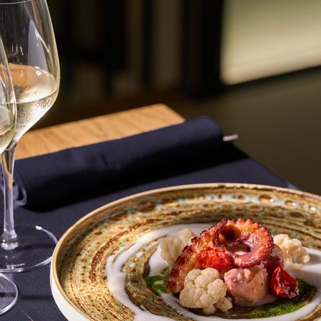 Piatto gourmet con tentacolo di polpo grigliato, cavolfiore e salsa verde, accompagnato da due calici di vino bianco su un tavolo scuro.