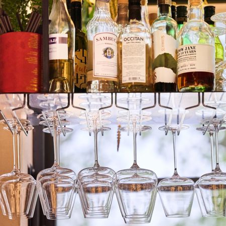 Dettaglio di un bancone bar con bottiglie di liquore sullo sfondo e calici di vino appesi a un porta bicchieri in primo piano.