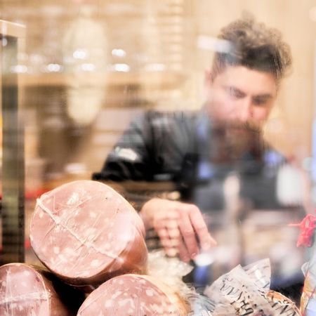 Salumi in primo piano nella vetrina refrigerata di un'enoteca, con lo chef al lavoro sullo sfondo.