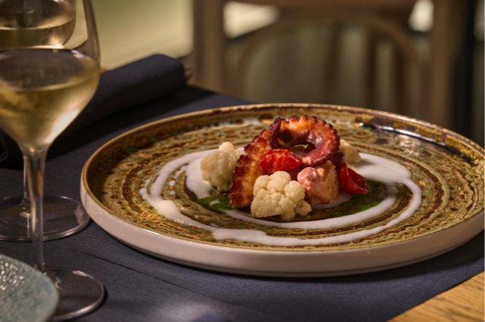 Piatto gourmet con tentacolo di polpo grigliato, cavolfiore e salsa verde, accompagnato da due calici di vino bianco su un tavolo scuro.