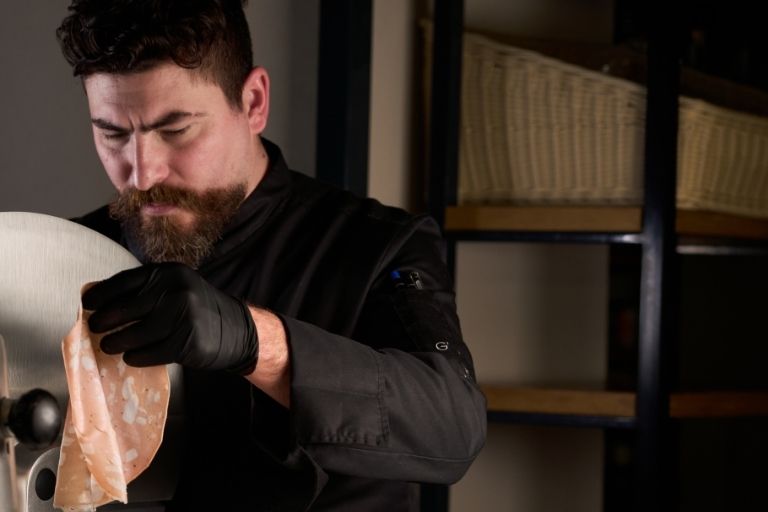 Chef con barba e guanti neri affetta finemente la mortadella con un'affettatrice professionale in un ambiente di cucina.